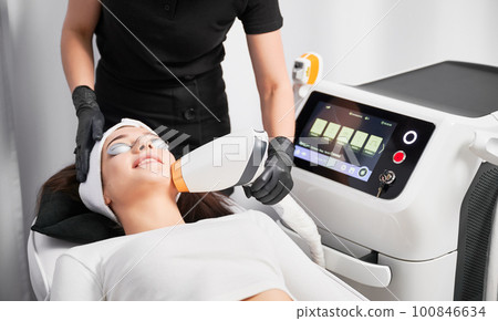Female patient in safety glasses lying near cosmetology equipment and having face lifting procedure. Doctor cosmetologist using laser scanner device while performing facial treatment in beauty salon. 100846634