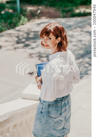 Business woman city street. A woman in a white shirt and blue denim skirt walks down the street holding a blue notebook in her hands. Summer city walks, travel Business woman city street. A woman in a white shirt and blue denim skirt walks down the street holding a blue notebook in her hands. Summer city walks, travel 100847048