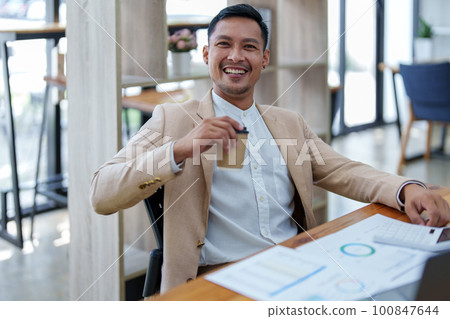 Portrait of a male business owner showing a happy smiling face as he has successfully invested his business using computers and financial budget documents at work. 100847644