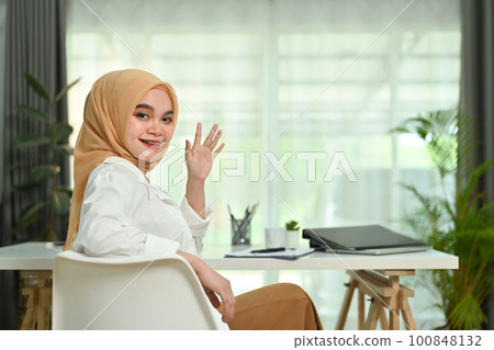 Positive asian Muslim woman worker in hijab waving hand at camera, greeting someone while sitting in modern office Positive asian Muslim woman worker in hijab waving hand at camera, greeting someone while sitting in modern office 100848132