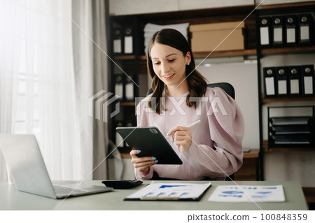Confident business expert attractive smiling young woman typing laptop ang holding digital tablet on desk in office. Confident business expert attractive smiling young woman typing laptop ang holding digital tablet on desk in office. 100848359