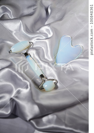 Blue jade face roller and Gua Sha scraping for beauty facial massage therapy on silver textured silk background. 100848361
