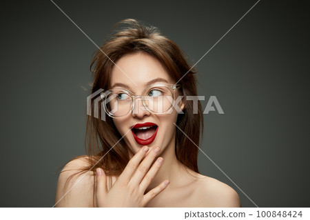 Emotive, young, beautiful girl with red lips makeup posing in stylish glasses against grey studio background 100848424