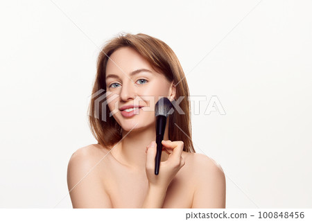 Young beautiful girl with well-kept clear face applying cosmetological product with brush against white background. Young beautiful girl with well-kept clear face applying cosmetological product with brush against white background. 100848456