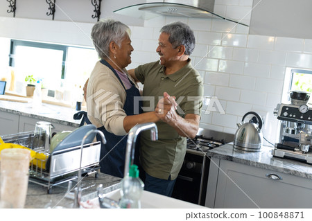 Side view of biracial senior couple holding hands and looking at each other while dancing in kitchen 100848871
