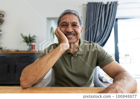 Portrait of smiling biracial senior man with hand on chin sitting at table at home, copy space 100848877