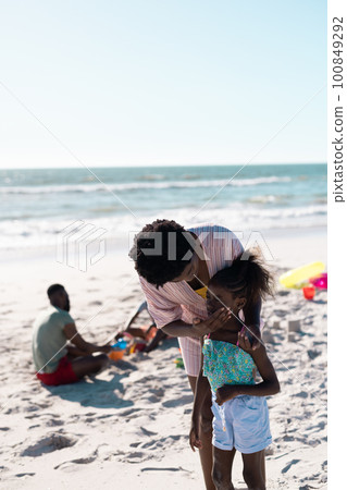 African american mother applying sunscreen on girl's face at beach against clear sky, copy space 100849292