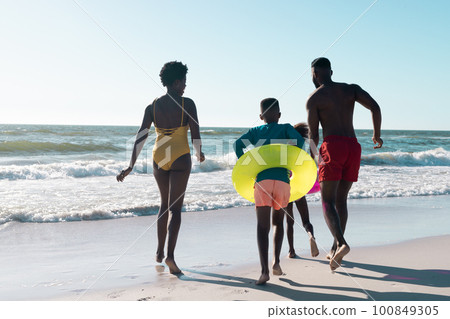 Rear view of cheerful african american parents and children with swimming floats running at seashore Rear view of cheerful african american parents and children with swimming floats running at seashore 100849305