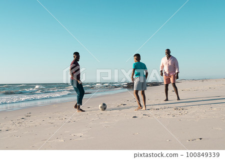 African american boy playing soccer with father and grandfather at beach against clear sky 100849339