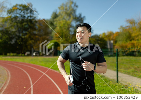 Young Asian male athlete running in the stadium, doing a morning run in headphones, holding a phone, listening to music. audiobook, podcast. 100852429