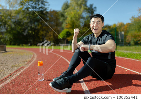 A young Asian man, an athlete sitting on a stadium treadmill. He looks at the smartwatch, fitness bracelet. Shows a gesture of victory and strength. Checks the results of running, training. A young Asian man, an athlete sitting on a stadium treadmill. He looks at the smartwatch, fitness bracelet. Shows a gesture of victory and strength. Checks the results of running, training. 100852441