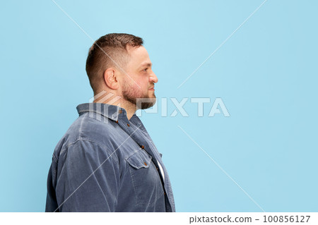 Profile view of one nice-looking man wearing jeans jacket standing with calm facial expression over blue background. Side view Profile view of one nice-looking man wearing jeans jacket standing with calm facial expression over blue background. Side view 100856127