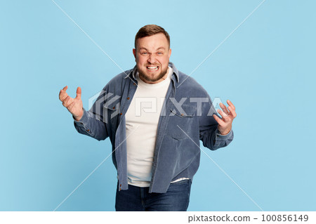 Portrait of angry bearded man with furious facial expression, screaming and spreading hands to sides over blue background 100856149