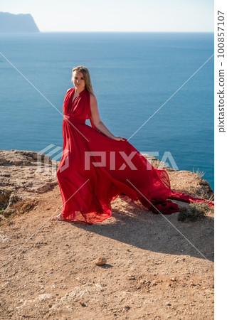 A woman in a red flying dress fluttering in the wind, against the backdrop of the sea. 100857107
