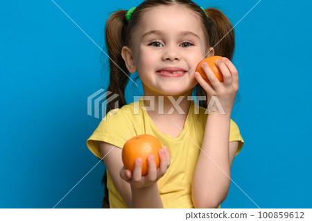 A small and smiling girl is holding two tangerines on a blue background. A small and smiling girl is holding two tangerines on a blue background. 100859612