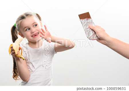 A child is offered chocolate and chooses a banana, on a white background a man's hand gives chocolate to a little girl who holds a banana in her hand. 100859631