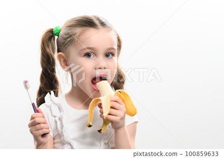 A little girl eats a banana and holds a toothbrush in her hands, studio photo on a white background, a banana helps against caries. 100859633