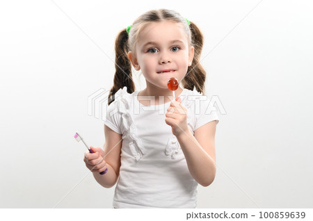little girl licks a lollipop and holds a toothbrush on a white background, copy space. little girl licks a lollipop and holds a toothbrush on a white background, copy space. 100859639