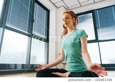 Low-angle view of calm wellness young woman meditating in lotus position sitting on floor with closed eyes at home enjoying meditation on background of window. Concept of healthy lifestyle. Low-angle view of calm wellness young woman meditating in lotus position sitting on floor with closed eyes at home enjoying meditation on background of window. Concept of healthy lifestyle. 100861586