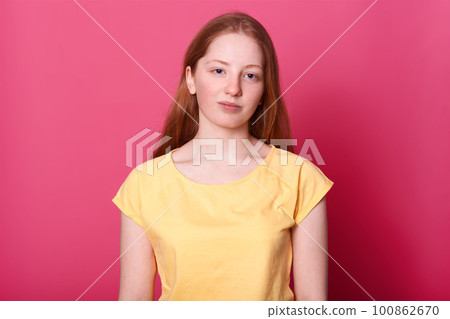 Calm schoolgirl of gentle appearance, naturally posing in studio alone, looks into camera isolated over pink background. Charming girl wears yellow t shirt, with straight brown hair. People concept. 100862670