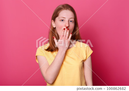 Attractive happy female touching lips. Beautiful brown haired girl keeps arm near her mouth and looks at camera. Model wears casual yellow t shirt poses isolated over pink background. Love concept. 100862682