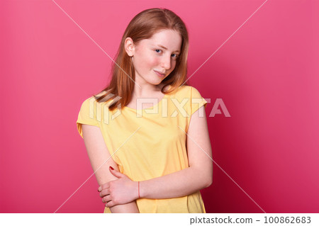 Charming magnetic positive girl poses isolated over bright pink background in studio, holding her arm, looking mysterious at camera. Talented sweet model wearing casual light yellow t shirt. 100862683