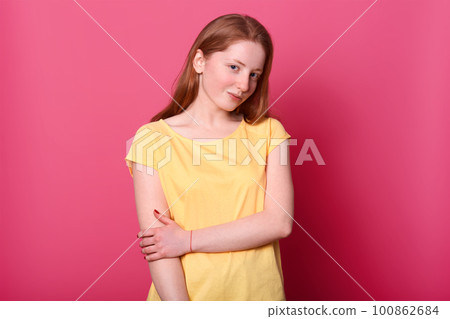 Romantic pretty young lady looking directly at camera, has shy facial expression, posing over pink background. Studio shot of girl with straight brown hair, touches her hand with another one. 100862684