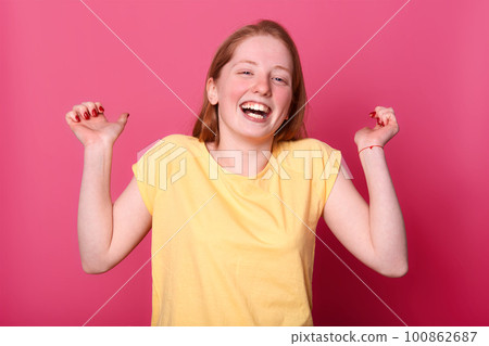 Studio shot of happy emotional young woman in bright yellow t shirt laughing sincerely, keeps hands up, hears funny joke, enjoys to spend time and to have fun with her friends. People emotions concept 100862687