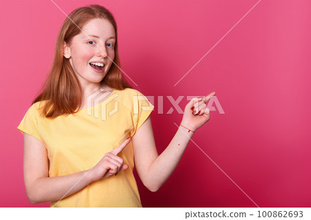Portrait of cute lovely charming cheerful teenager girl showing something pointing aside with fore fingers, female stands smiling over studio wall. Blank space for promotion text or advertisment. Portrait of cute lovely charming cheerful teenager girl showing something pointing aside with fore fingers, female stands smiling over studio wall. Blank space for promotion text or advertisment. 100862693
