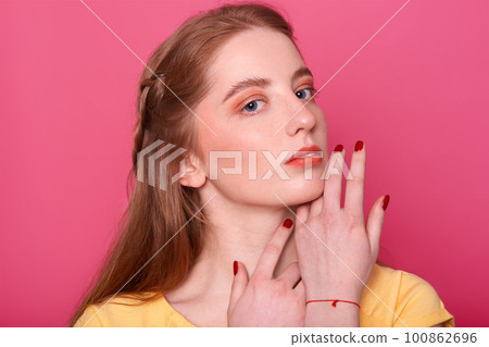 Gentle woman with bright makeup, straight hair with red shade, has manicured hand. Model in yellow shirt, keeps hands near chin, looks at camera, has mysteriously facial expression. Beauty concept. 100862696