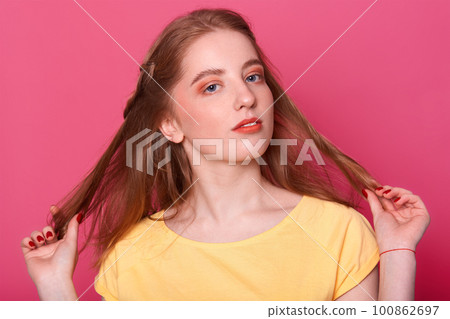 Closeup portrait of beautiful young woman holds her straight long shiny brown hair. Attractive girl witn bright makeup, wears yellow t shirt, looks mysterious, poses isolated on rose background. 100862697