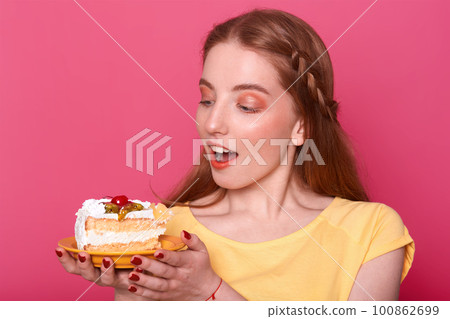 Attractive young woman with opened mouth holds plate with piece of delicious cake in hands. Brown haired lady with red manicure poses in studio isolated over pink background. Sweet food concept. Attractive young woman with opened mouth holds plate with piece of delicious cake in hands. Brown haired lady with red manicure poses in studio isolated over pink background. Sweet food concept. 100862699