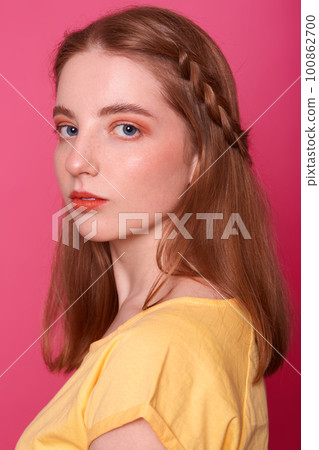 Close up shot of adorable Caucasian female with straight hair, dressed in yellow t shirt, gently looking at camera, has confident facial expressions, poses in studio isolated over pink background. Close up shot of adorable Caucasian female with straight hair, dressed in yellow t shirt, gently looking at camera, has confident facial expressions, poses in studio isolated over pink background. 100862700