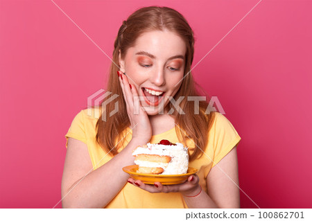 Close up portrait of satisfied pleased girl with light brown hair, holds huge piece of tasty cake, keeps her mouth open, full of enjoyment, dressed in casual yellow t shirt, poses in photo studio. 100862701