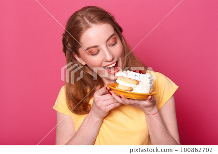 Photo of young female in casual yellow t shirt, ready for eating huge piece of cake, looks at tasty pastry, isolated on rose background, studio portrait. Culinary, people and lifestyle concept. Photo of young female in casual yellow t shirt, ready for eating huge piece of cake, looks at tasty pastry, isolated on rose background, studio portrait. Culinary, people and lifestyle concept. 100862702