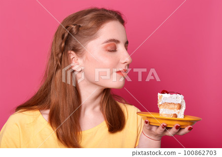 Horizontal shot of good looking young female, model sniffs tasty dessert, concentared at huge piece of cake, wants to eat delicious pastry, poses in photo studio isolated over pink background. Horizontal shot of good looking young female, model sniffs tasty dessert, concentared at huge piece of cake, wants to eat delicious pastry, poses in photo studio isolated over pink background. 100862703