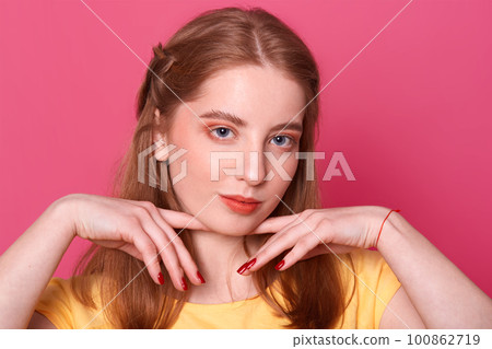 Romantic tender young lady holds fingers with red manicure near cute fresh face with make up posing over dark pink background in studio, having red thread on her wrist, looks sweet and pure. Romantic tender young lady holds fingers with red manicure near cute fresh face with make up posing over dark pink background in studio, having red thread on her wrist, looks sweet and pure. 100862719