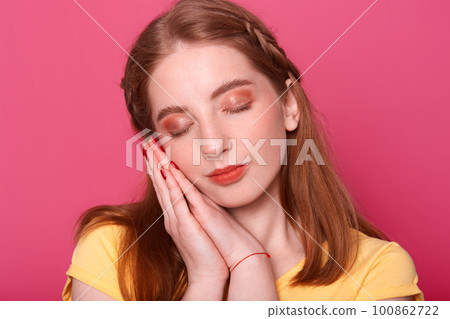 Indoor studio shot of romantic pretty young lady with closed eyes and hands near face, going to have nap, looks sweet. Attractive model poses isolated over bright pink background. Youth concept. 100862722