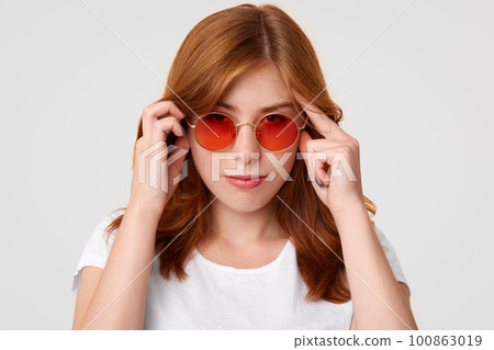 Concentrated young European female with serious expression, keeps hands on temples, tries to focus on something, wears red sunglasses and white casual t shirt, isolated over studio background Concentrated young European female with serious expression, keeps hands on temples, tries to focus on something, wears red sunglasses and white casual t shirt, isolated over studio background 100863019