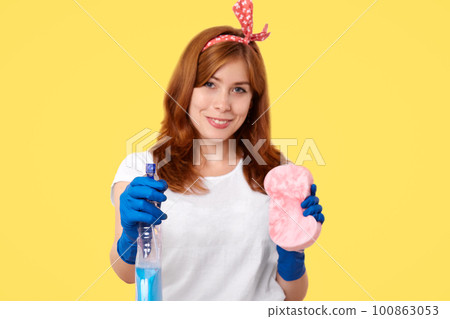 People and housekeeping concept. Adorable smiling female housewife in casual clothes, does house duties, cleans in room, holds bottle with detergent and sponge, stands over yellow background 100863053