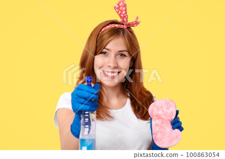 Lovely young housemaid in headband, rubber protective gloves, uses washing spray and sponge for cleaning mirror, smiles positively, isolated over yellow background, rejoices finishing housework Lovely young housemaid in headband, rubber protective gloves, uses washing spray and sponge for cleaning mirror, smiles positively, isolated over yellow background, rejoices finishing housework 100863054