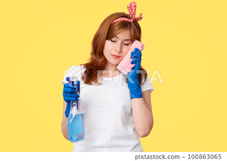 Sleepy female leans at sponge, being tired after tudying up, holds bottle with detergent, wears headband, dressed in casual clothes, isolated over yellow background. People and cleaning concept Sleepy female leans at sponge, being tired after tudying up, holds bottle with detergent, wears headband, dressed in casual clothes, isolated over yellow background. People and cleaning concept 100863065