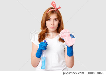 Serious housemaid looks with self confident expression, has ginger hair, wears rubber gloves, holds spray and sponge for cleaning furniture, isolated over white background. Housekeeping concept Serious housemaid looks with self confident expression, has ginger hair, wears rubber gloves, holds spray and sponge for cleaning furniture, isolated over white background. Housekeeping concept 100863066