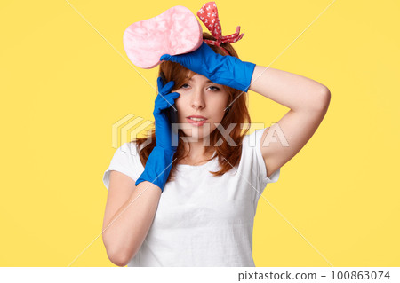 Cleaning and disinfection concept. Tired busy housewife feels overworked after spring cleaning, holds mop and rubs forehead, discusses housework with friend via smart phone. Domestic routine 100863074