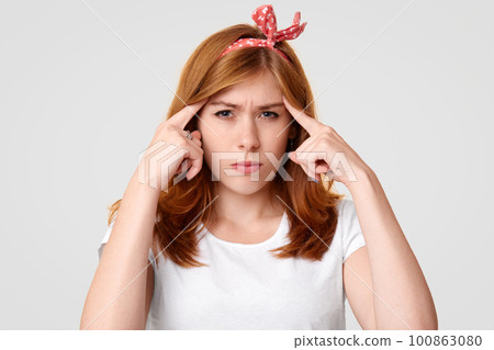 Serious concentrated female with displeased expression, keeps fore fingers on temples, tries to focus on some information, recollects something in mind, wears headband and white casual t shirt 100863080
