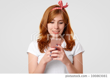 Isolated shot of beautiful smiling ginger female with charming smile, uses modern mobile phone, messages with friends in social networks, recreats with technology, poses in studio. Lifestyle concept 100863081