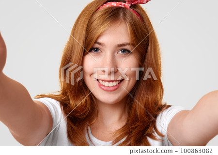 Adorable satisfied female model makes selfie with unrecongnizable device, stretches hands, has lovely expression, isolated over white background. Close up portrait of happy woman in retro style 100863082