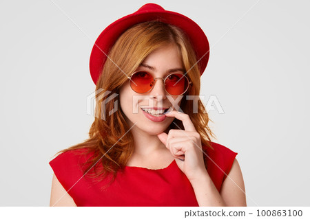 Glamour attractive young female keeps finger near mouth, smiles broadly, shows white teeth, dressed in red clothes, wears stylish sunglasses, being in high spirit after celebrating her birthday Glamour attractive young female keeps finger near mouth, smiles broadly, shows white teeth, dressed in red clothes, wears stylish sunglasses, being in high spirit after celebrating her birthday 100863100