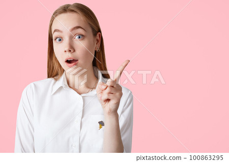 Lovely young female remembers about important meeting, keeps fore finger raised or gets idea, wears elegant white shirt, isolated on pink background with blank copy space for your advertisement 100863295