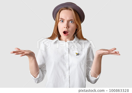 Photo of indignant young woman gestures angrily, opens mouth widely, wears elegant shirt and headgear, has puzzled displeased expression, isolated over white background. Negative feeling concept Photo of indignant young woman gestures angrily, opens mouth widely, wears elegant shirt and headgear, has puzzled displeased expression, isolated over white background. Negative feeling concept 100863328
